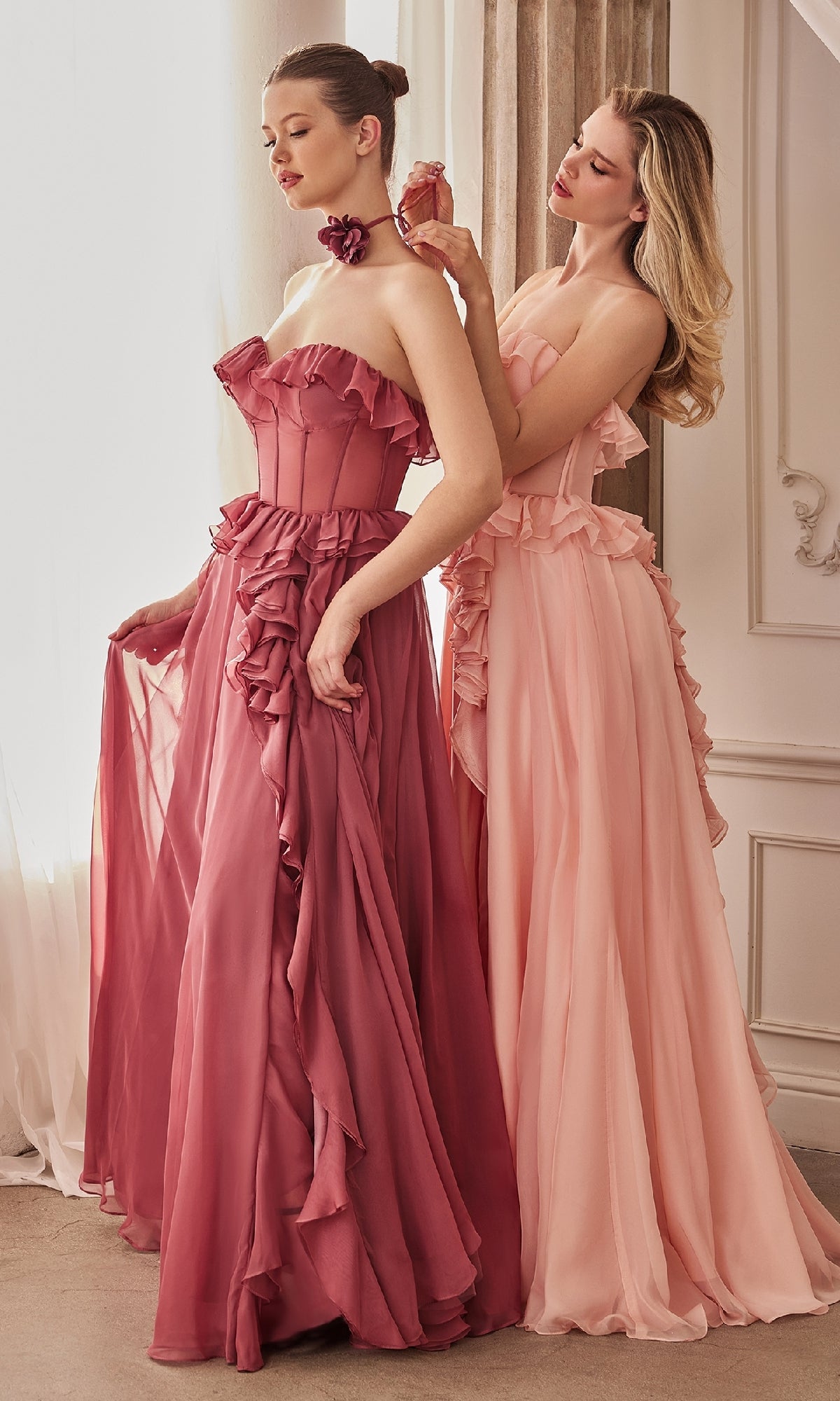 Two women in elegant gowns—one wearing the Andrea and Leo A1341 Strapless Ruffled Prom Dress in deep rose, the other in pale pink. In a softly lit, ornate room, one sprays perfume on the other.